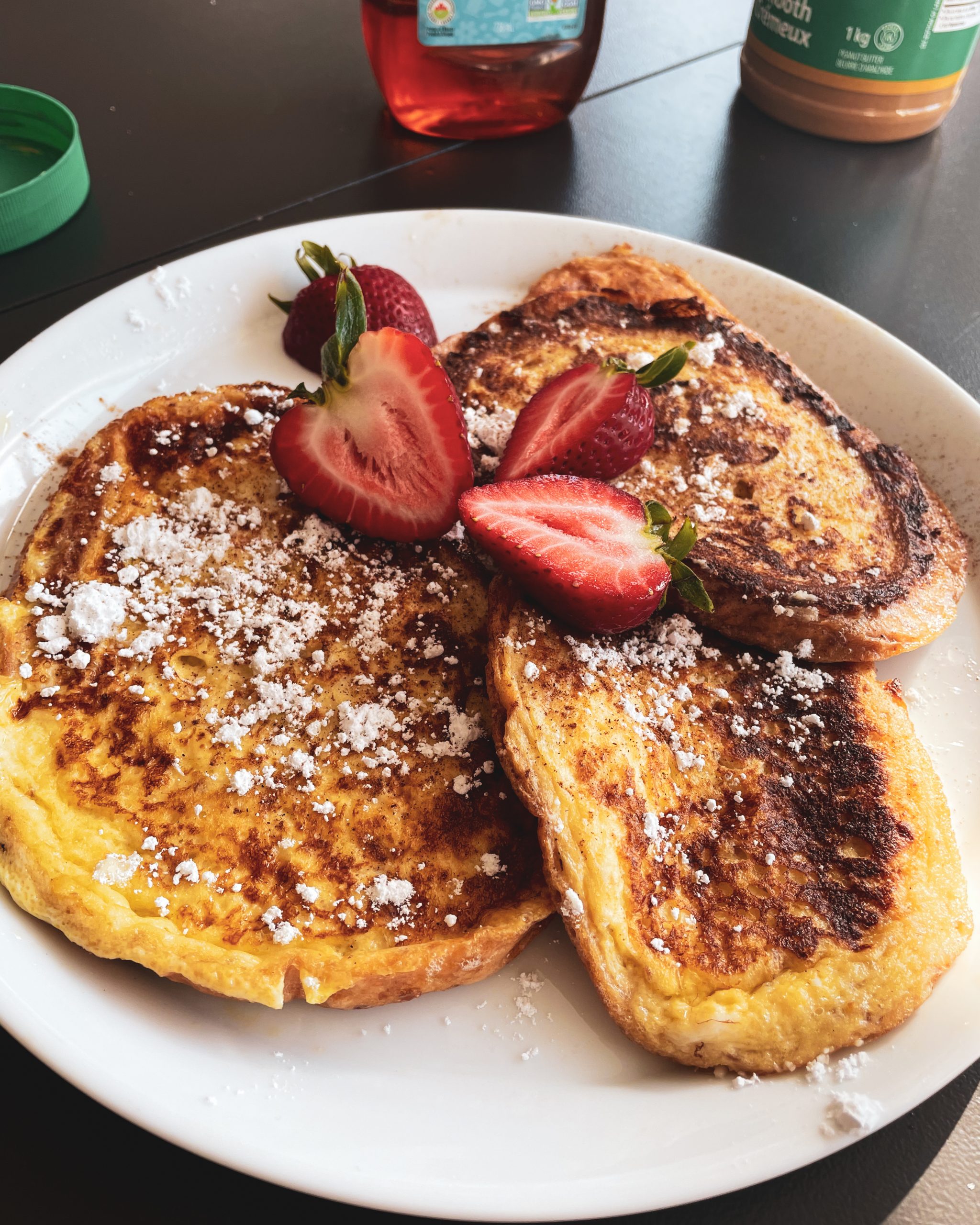 White plate with french toast sprinkled with powdered sugar and cinnamon and halved fresh straweberries